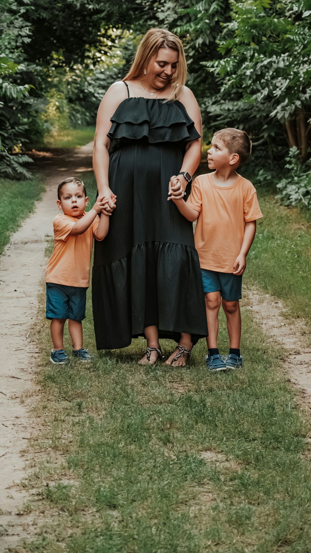 Pregnant woman in black dress holds hands with two young boys in matching orange shirts on a tree-lined path.