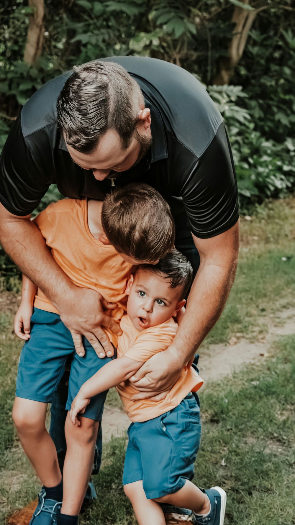 A father playfully wraps his arms around two young boys in matching orange shirts outdoors.