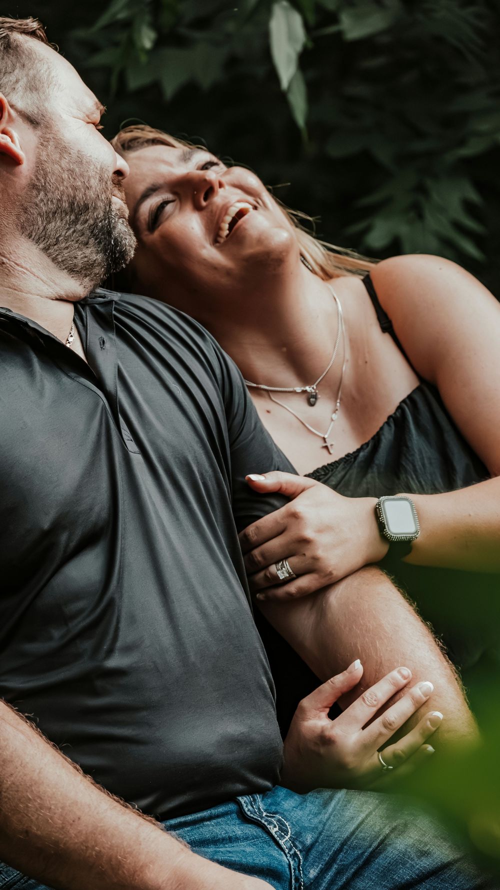 Happy couple embracing outdoors, woman laughing joyfully as man kisses her cheek among green foliage.