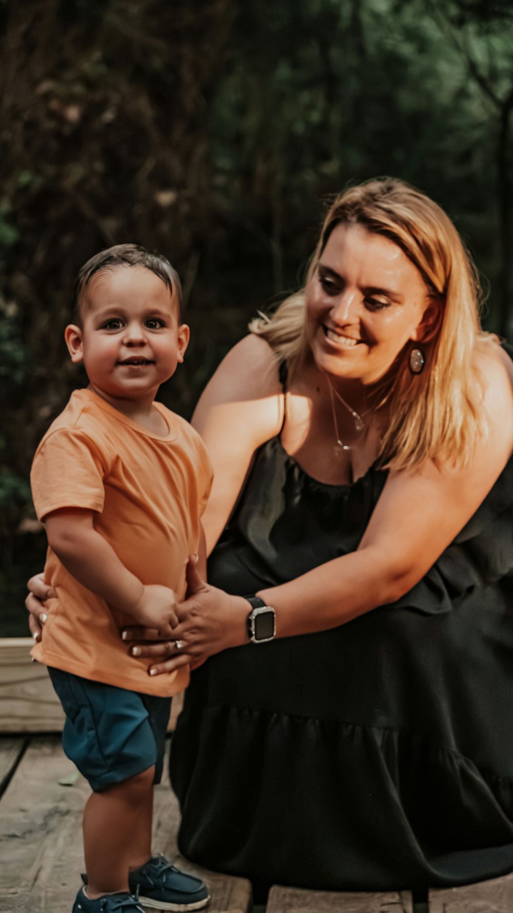 A smiling blonde woman in a black dress holds hands with a toddler boy in an orange shirt outdoors.