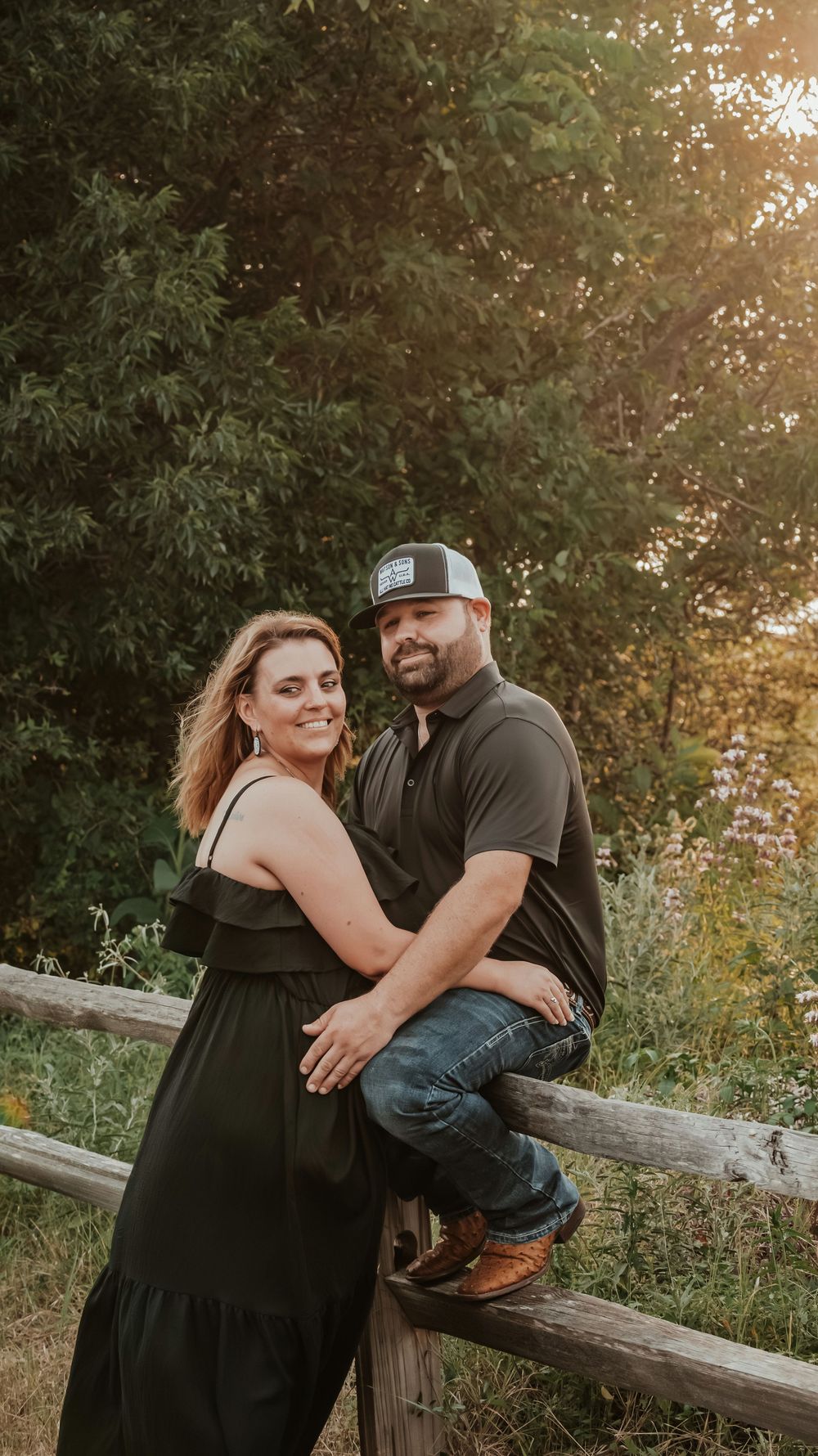 A couple in black outfits poses together by a wooden fence surrounded by lush greenery at sunset.