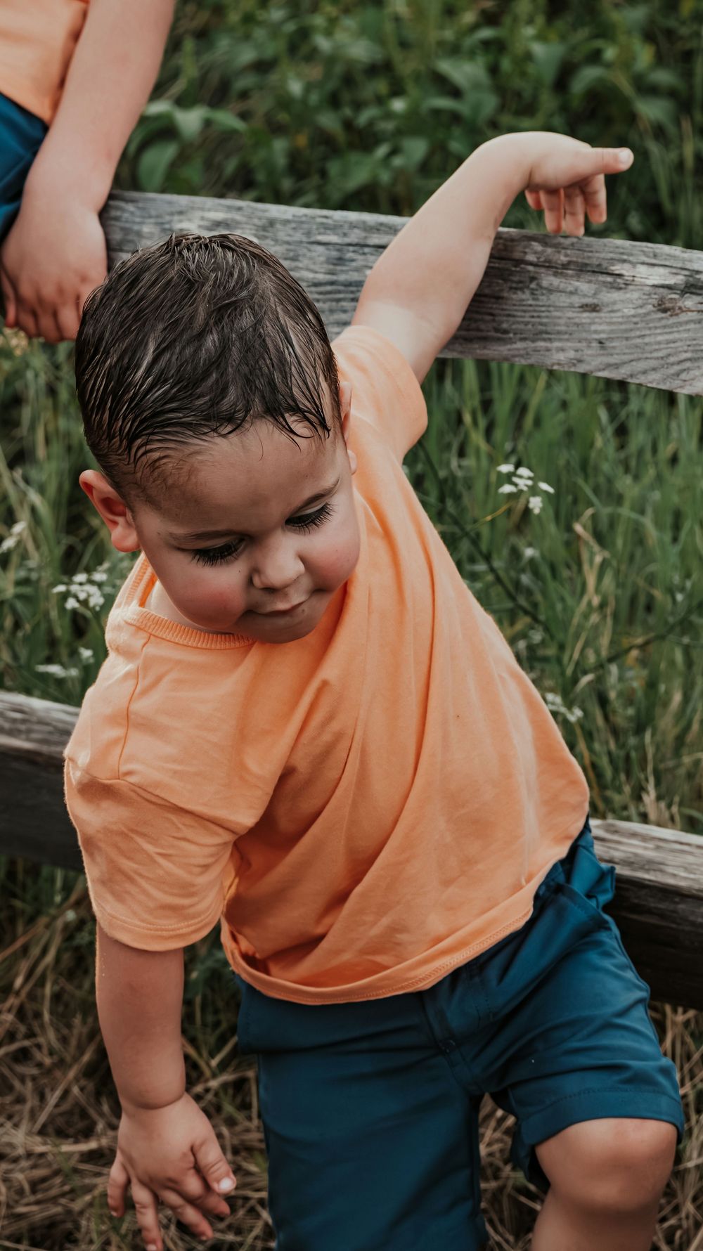 A young boy in an orange shirt and blue shorts plays near a wooden fence in a grassy field.