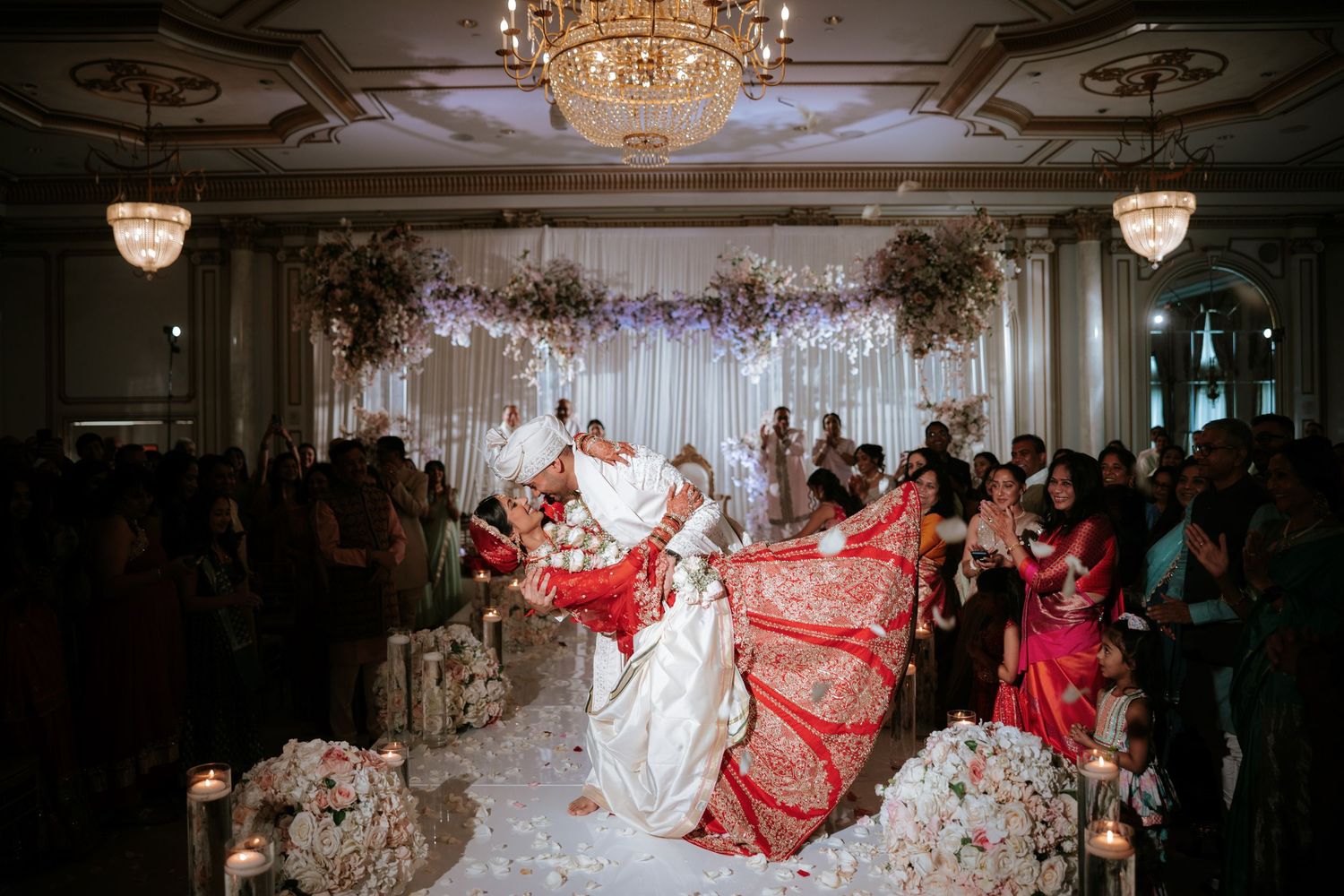 Bride in white gown and red dupatta during Indian wedding ceremony in elegant ballroom with floral arch and chandelier.