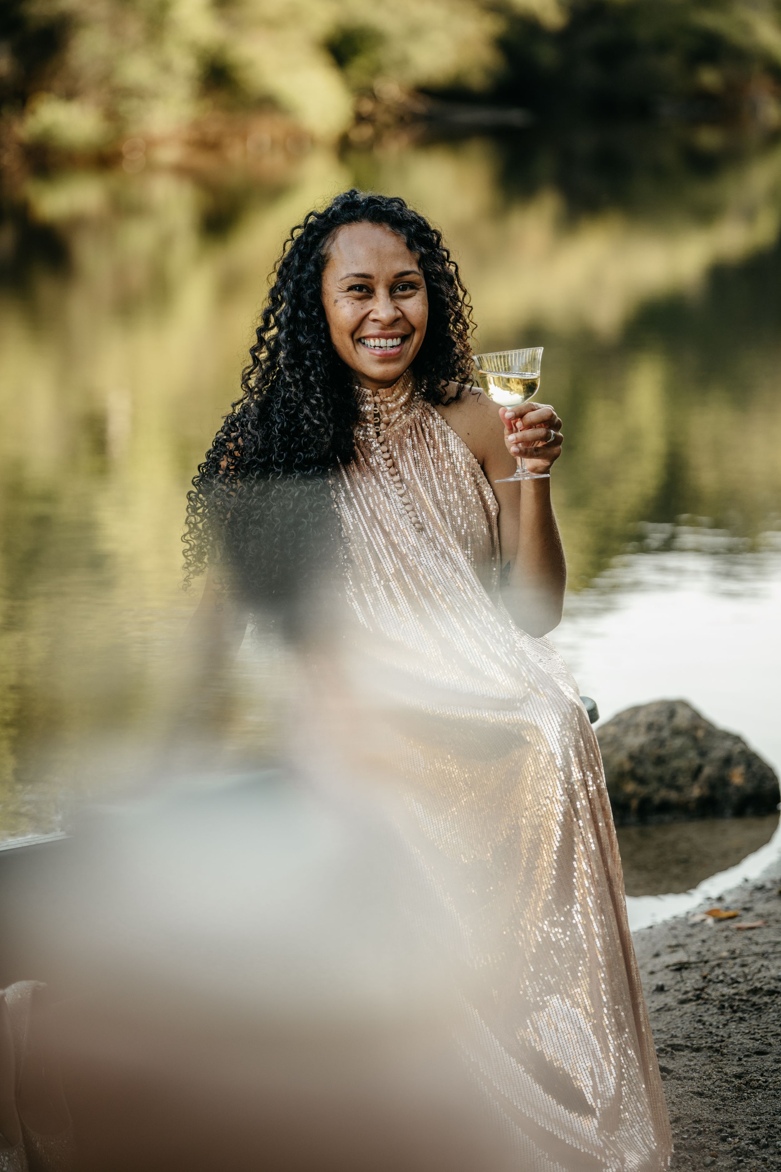 Smiling woman in a sequin dress holding a glass of white wine while sitting by a reflective river at golden hour.