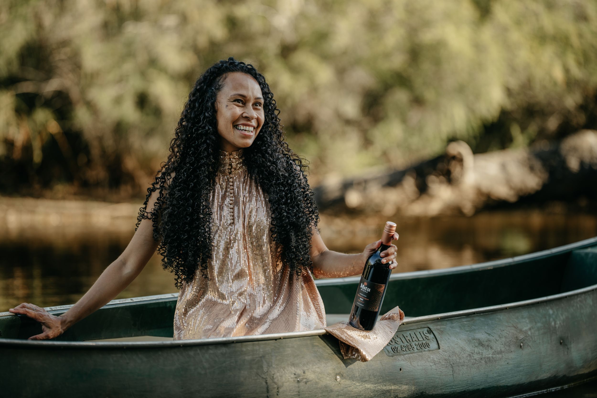 Smiling woman with curly hair holding a wine bottle while sitting in a canoe outdoors.
