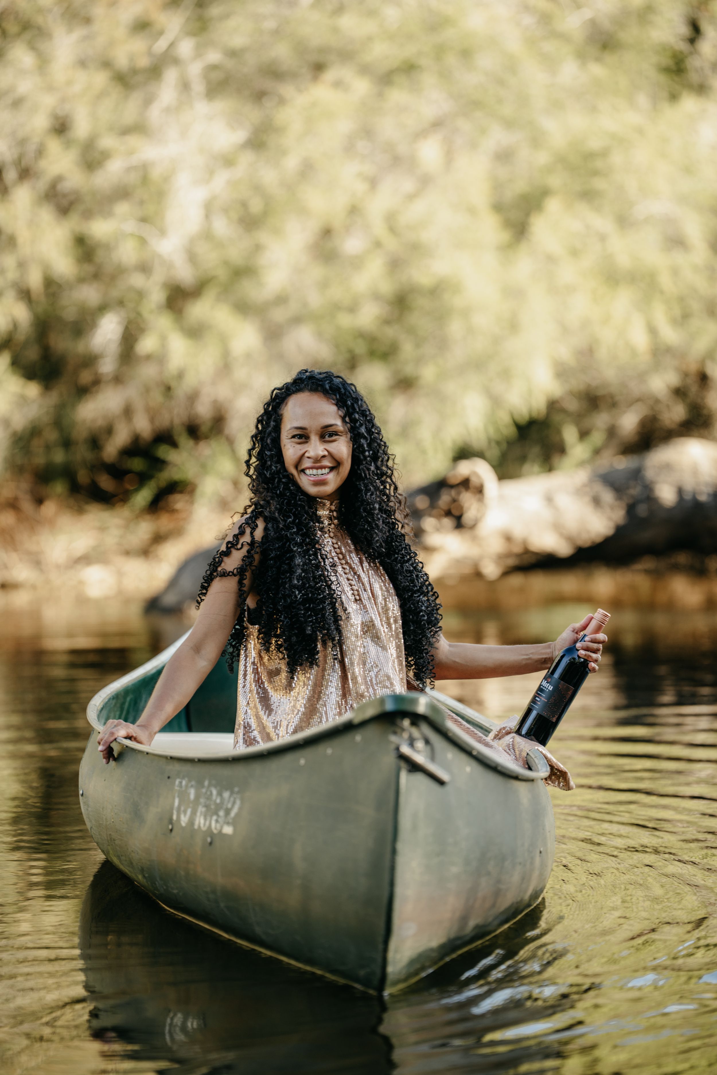Smiling woman with curly hair holding a wine bottle while sitting in a canoe on a calm river surrounded by trees.