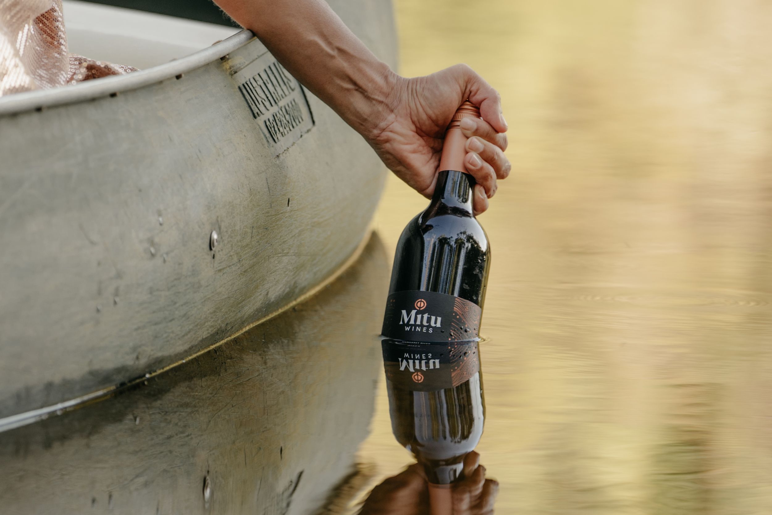 Close-up of a hand holding a dark wine bottle over water beside a canoe, with a reflection visible below.