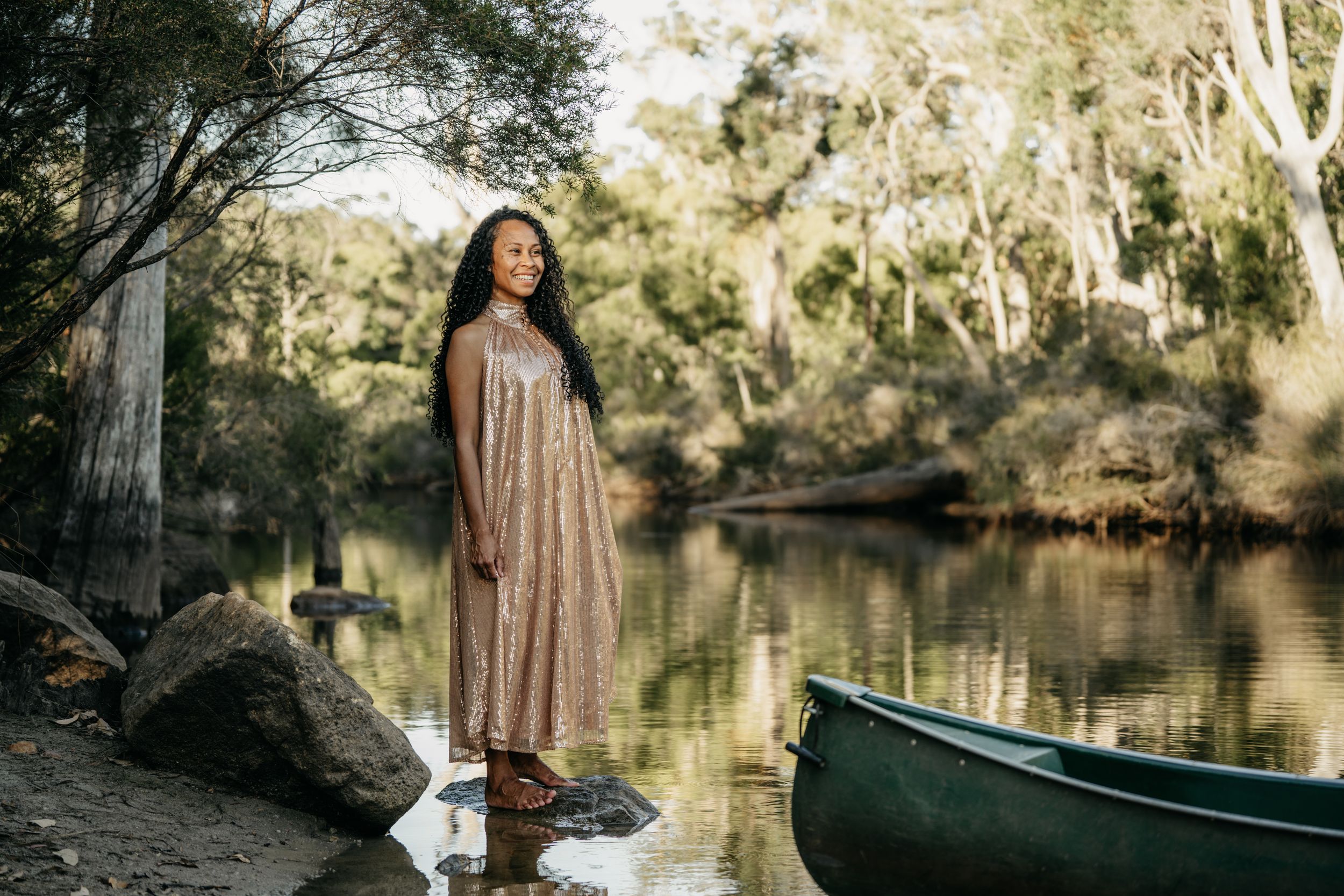 Woman in a rose gold sequin dress standing barefoot by a river next to a green canoe among eucalyptus trees.