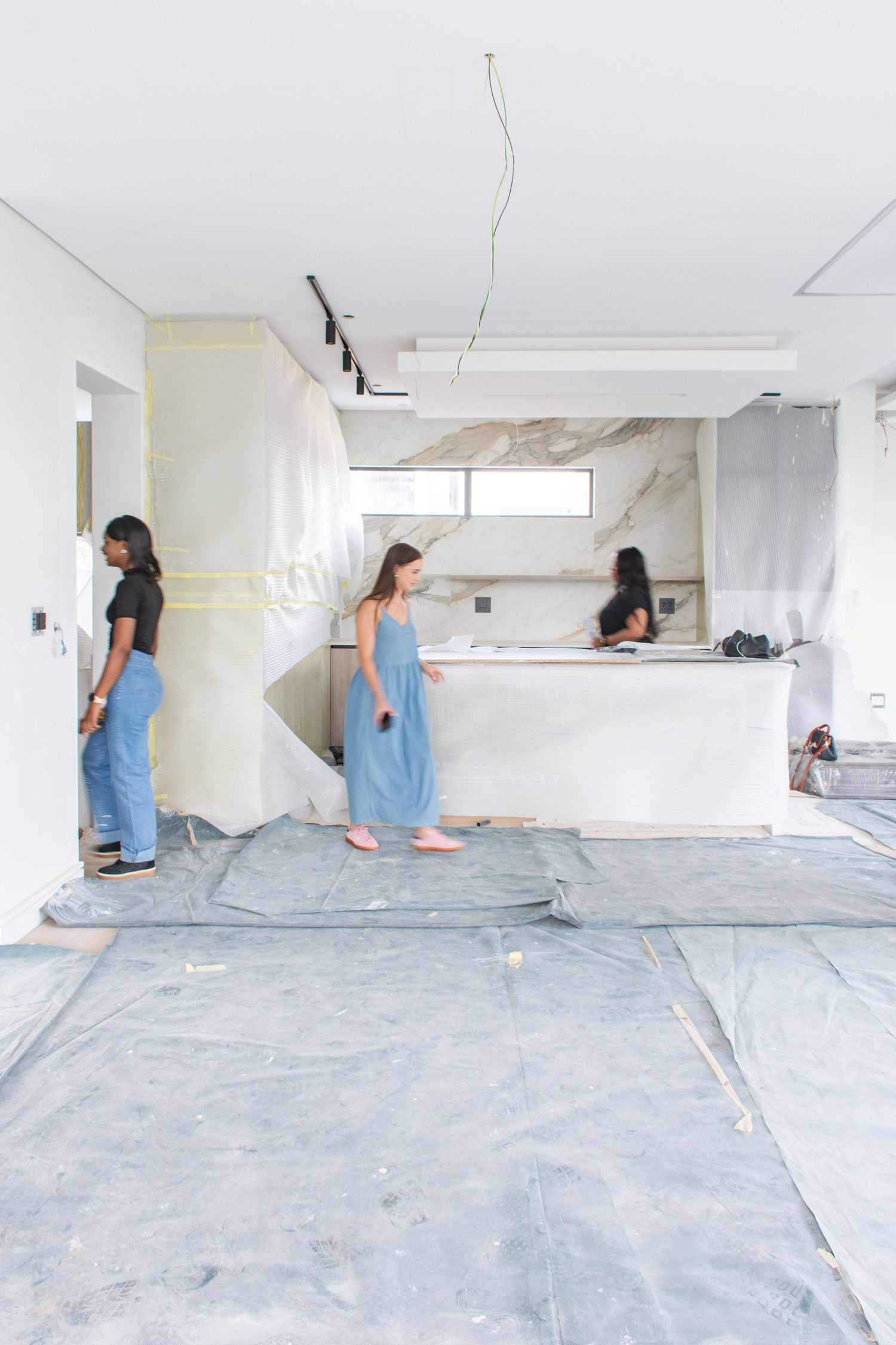 Three women in a modern kitchen under renovation, with marble walls, drop cloths on floor, and track lighting above.