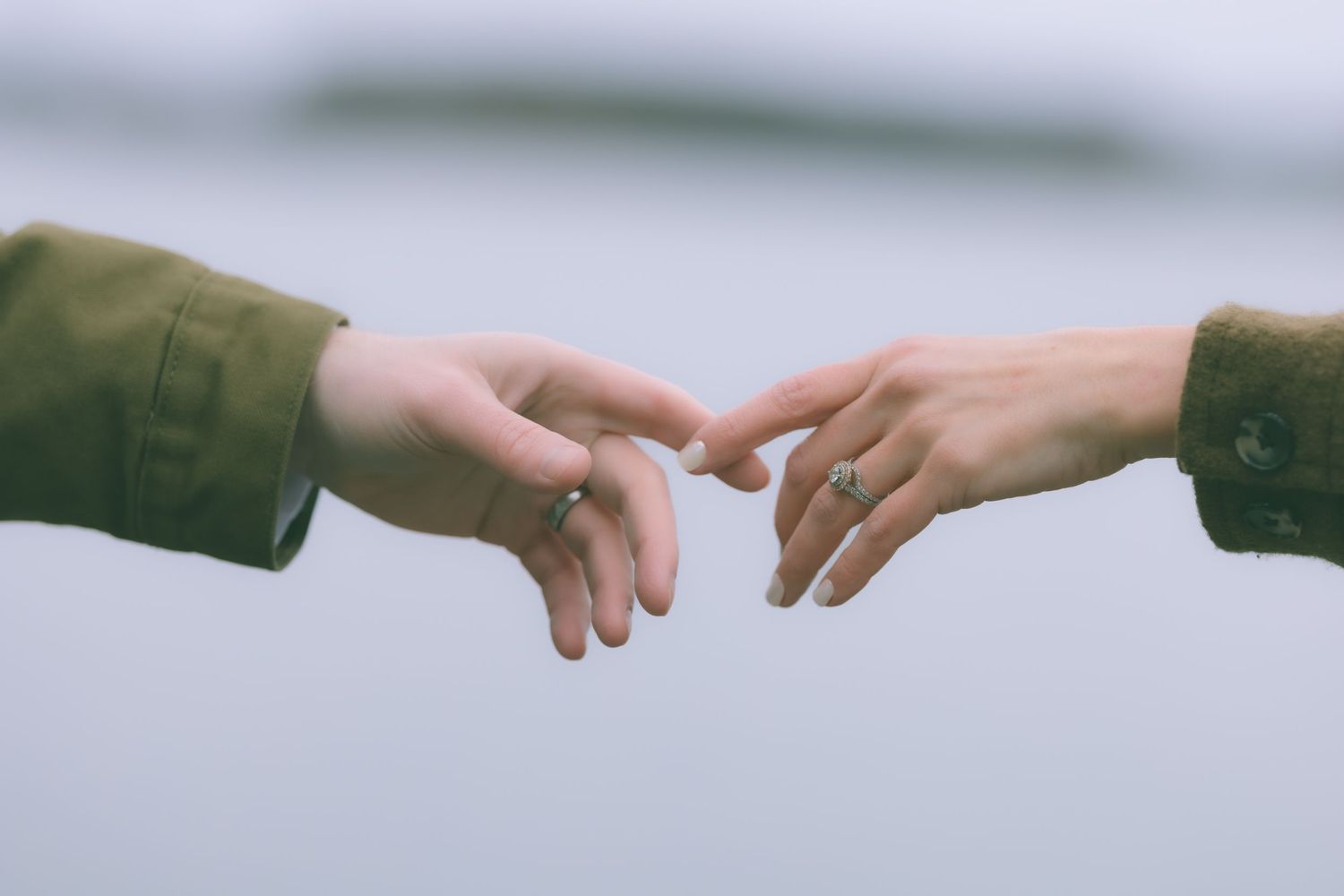 Two hands reaching toward each other with rings, nearly touching against blurred gray background.