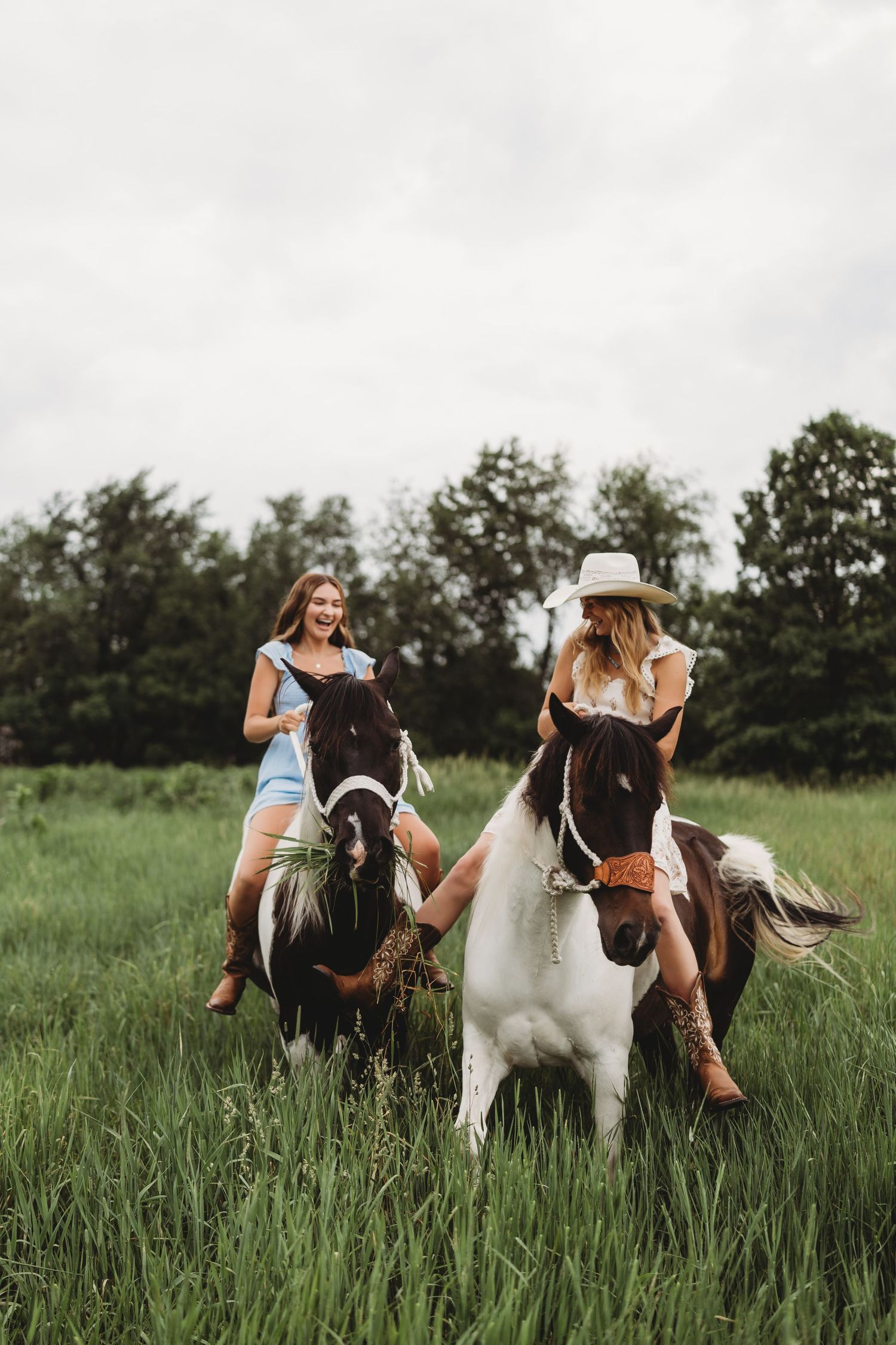 Two women riding painted horses through a lush green field with trees in the background on a cloudy day.