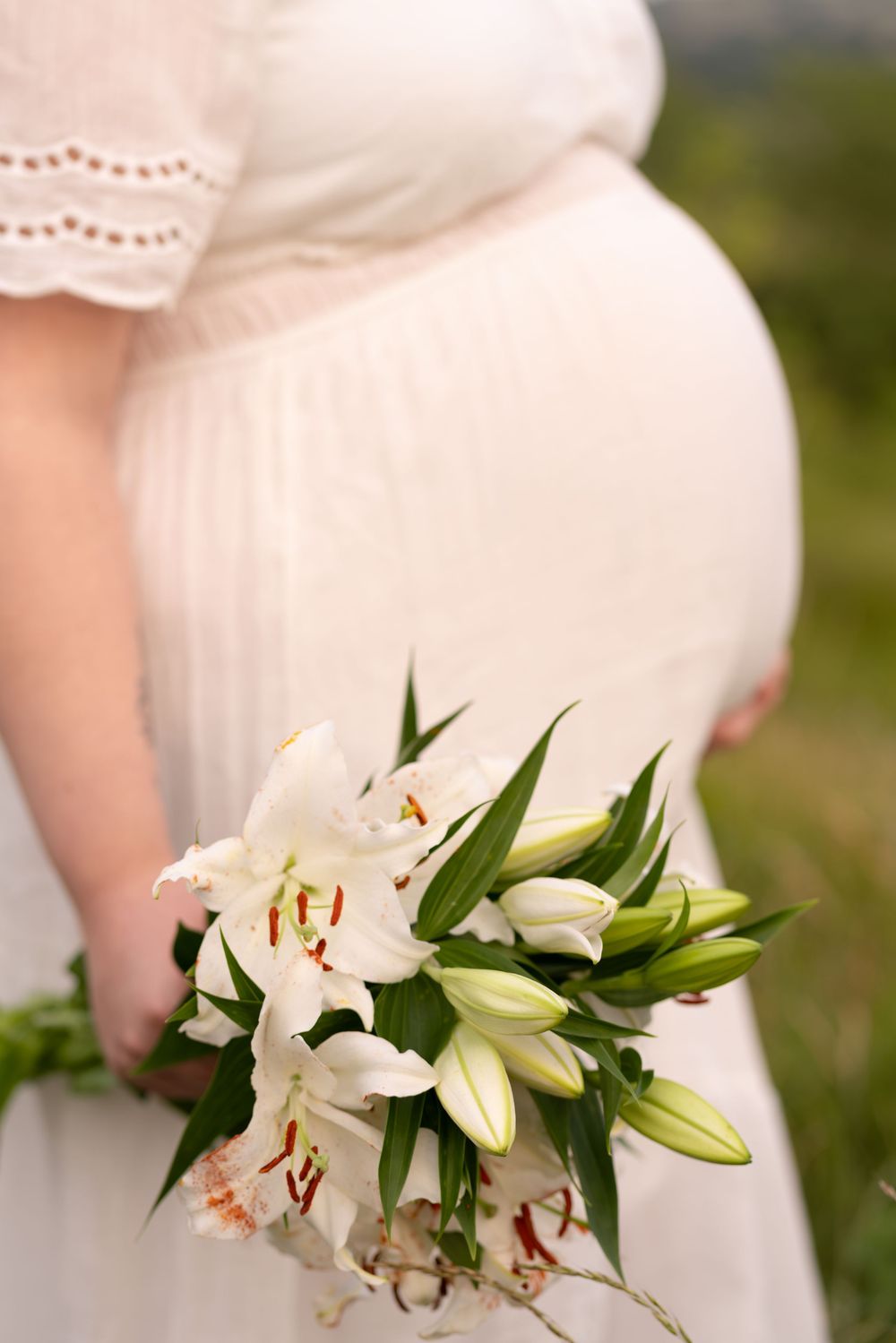 Pregnant woman in white dress holding bouquet of white lilies outdoors, showcasing maternity elegance.