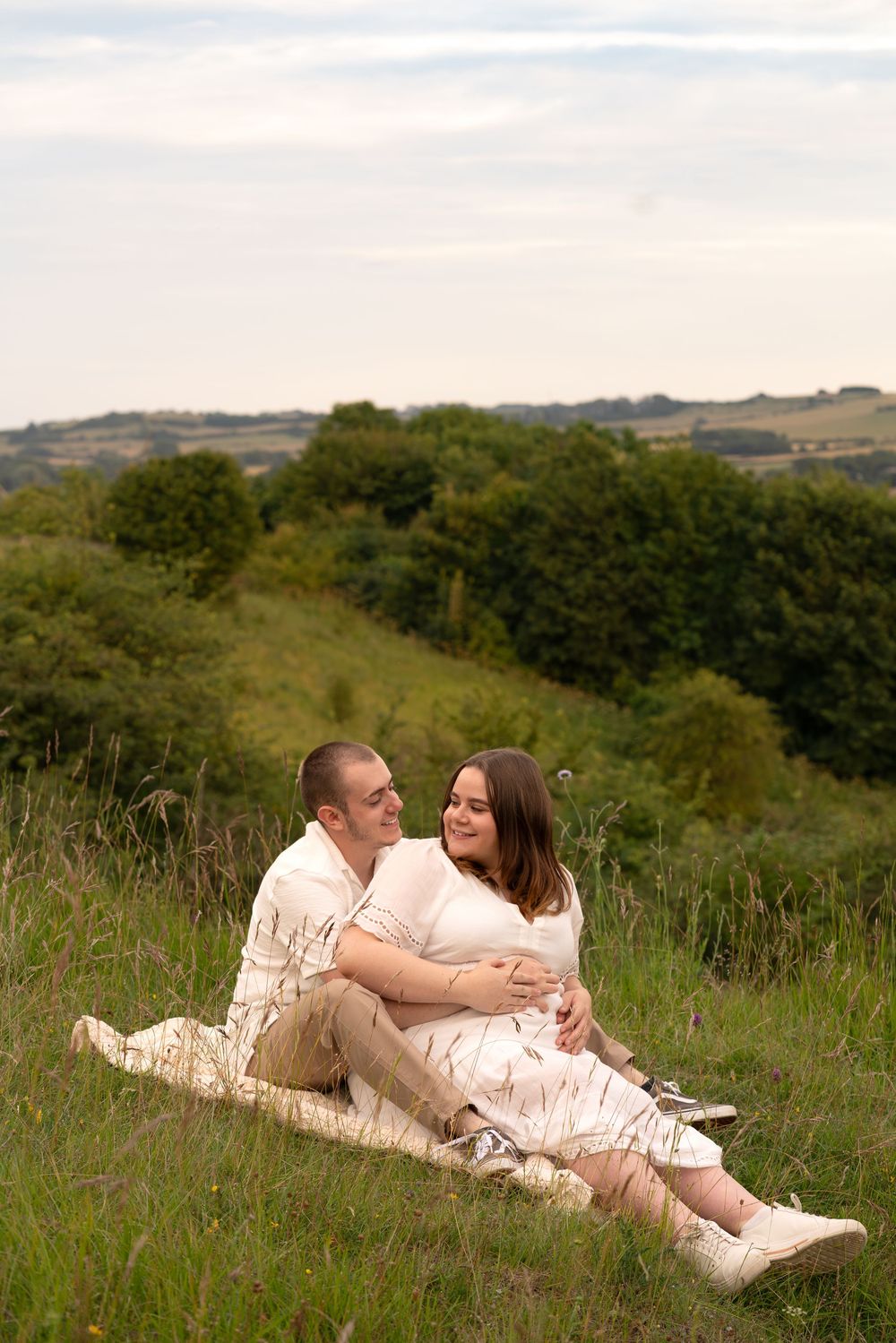 A couple sits together on a blanket in a lush green field, smiling and embracing on a cloudy day.