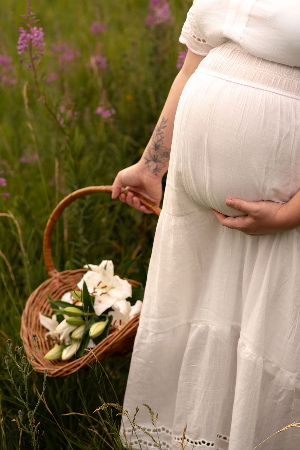Pregnant woman in white dress holding wicker basket with white lilies in a wildflower meadow.