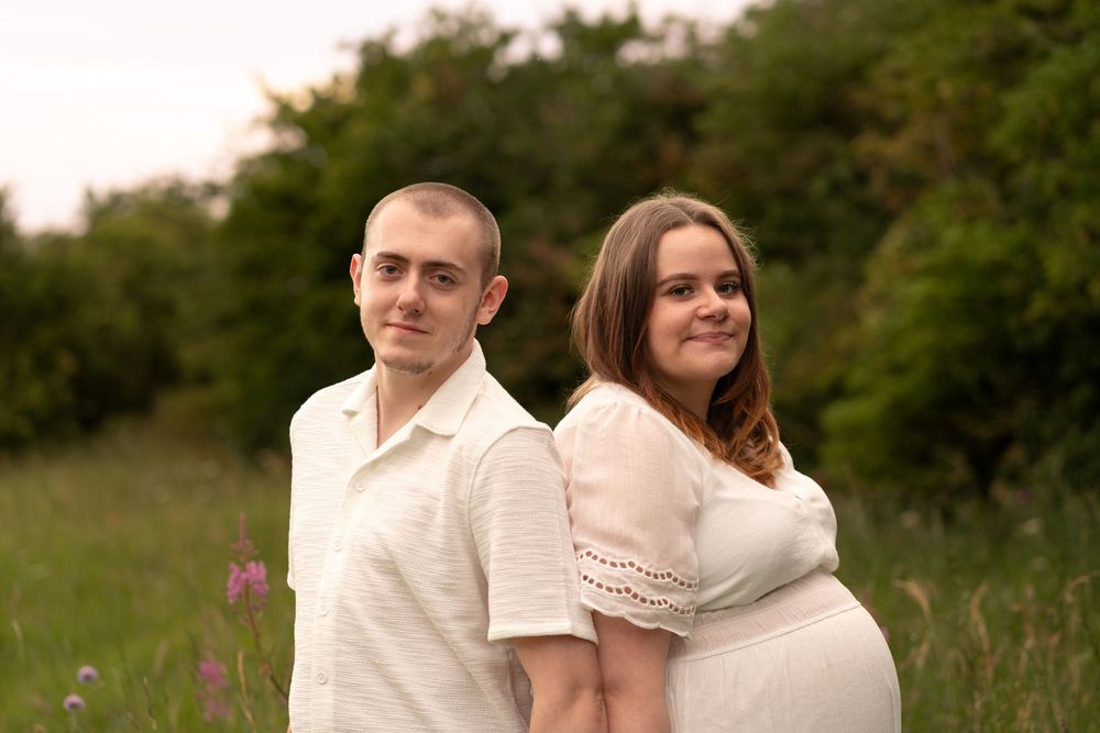 Couple posing back-to-back in a green meadow, woman in white dress showing baby bump, maternity photo session outdoors.