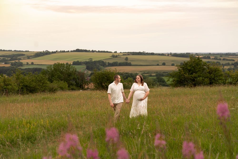 Pregnant couple holding hands in a lush green meadow with rolling hills at sunset.