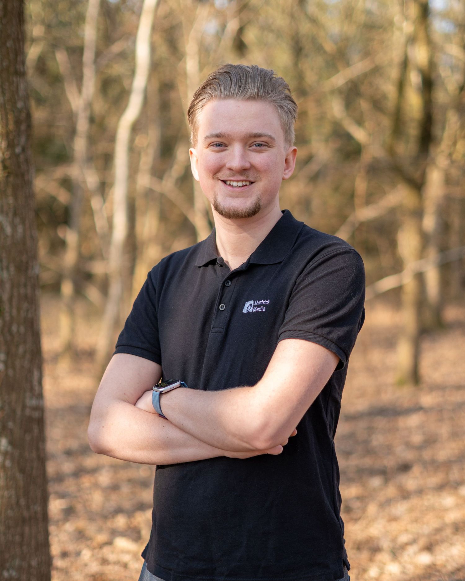 Young man with blonde hair smiling, arms crossed, wearing black polo shirt, standing outdoors in a wooded area.