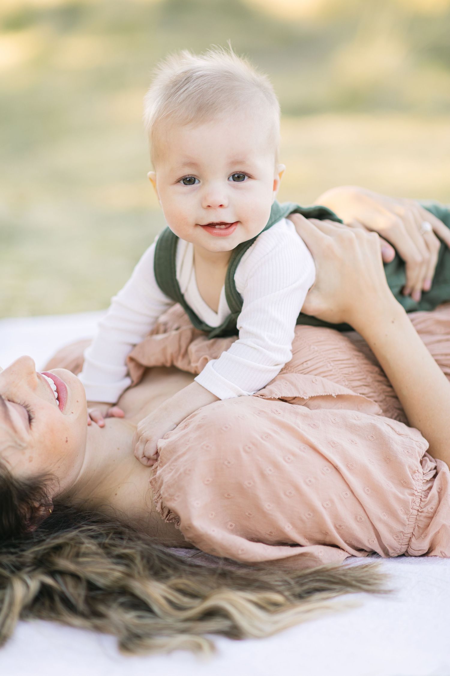 A laughing mother lies on a blanket outdoors while her smiling baby boy in green overalls plays on her chest.
