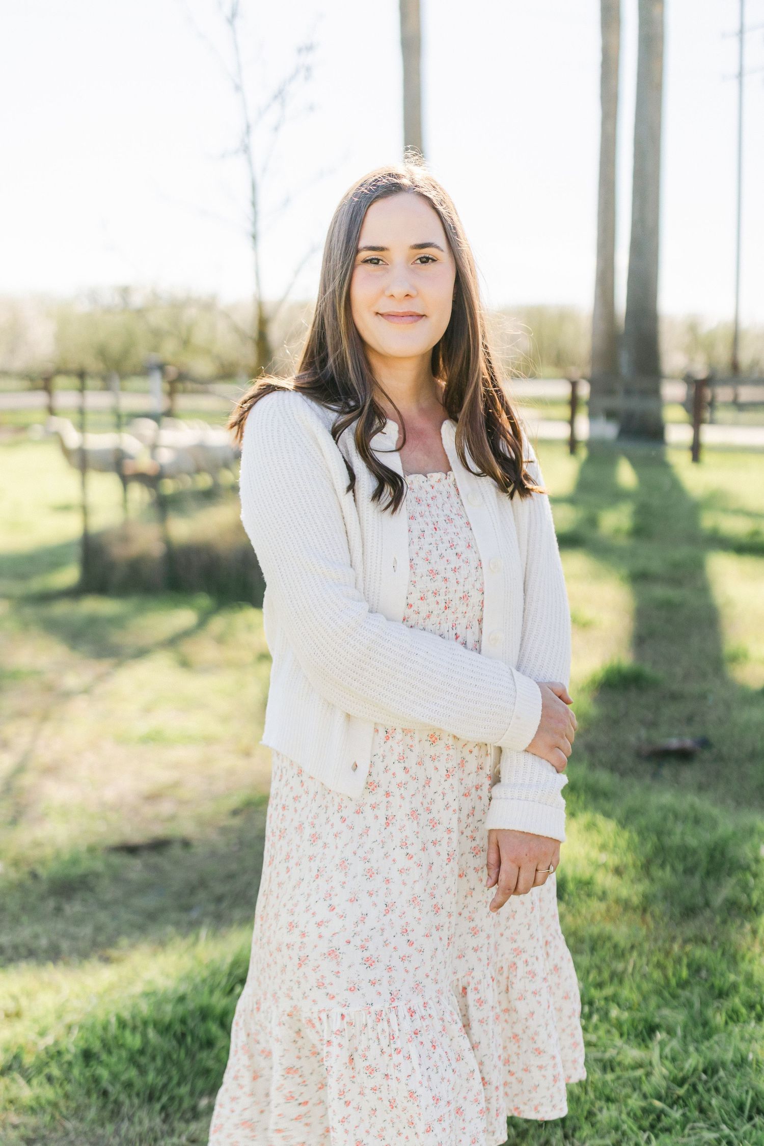 Woman in floral dress and white cardigan standing outdoors in golden hour light.