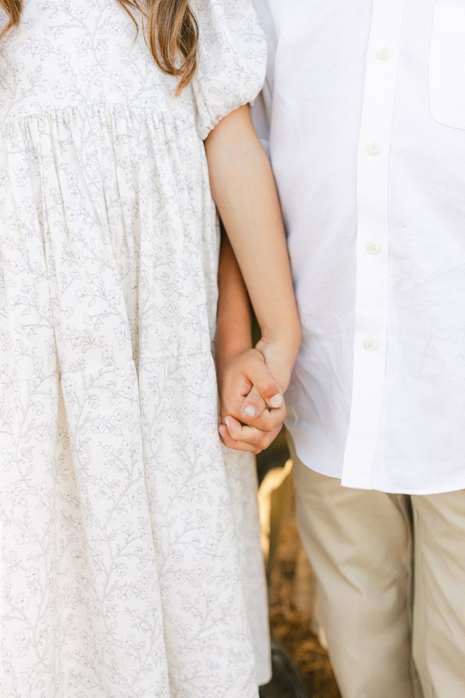 Two children holding hands, girl in white floral dress and boy in white shirt with khaki pants.