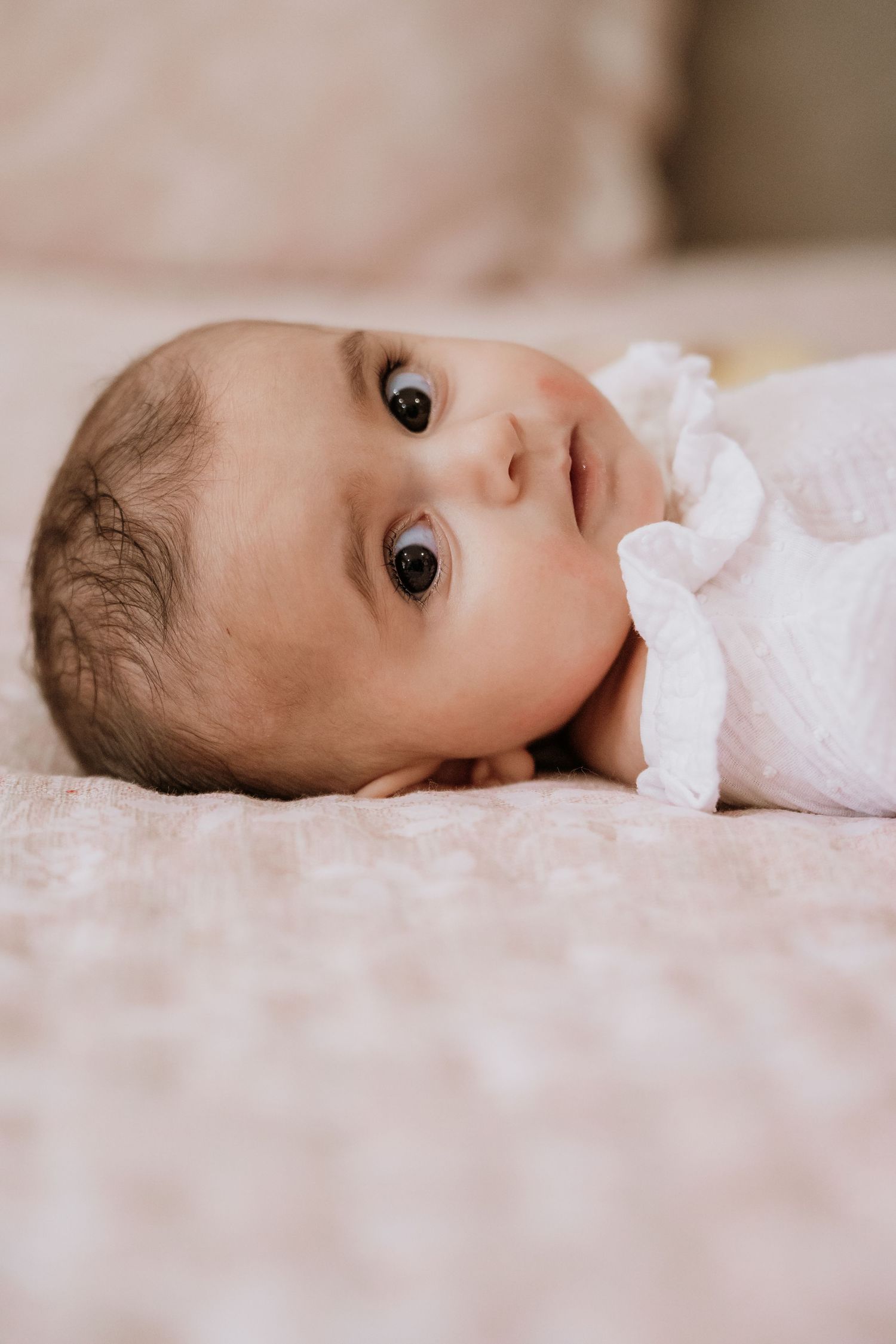 Newborn baby with dark eyes lying on a soft pink blanket wearing a white outfit.