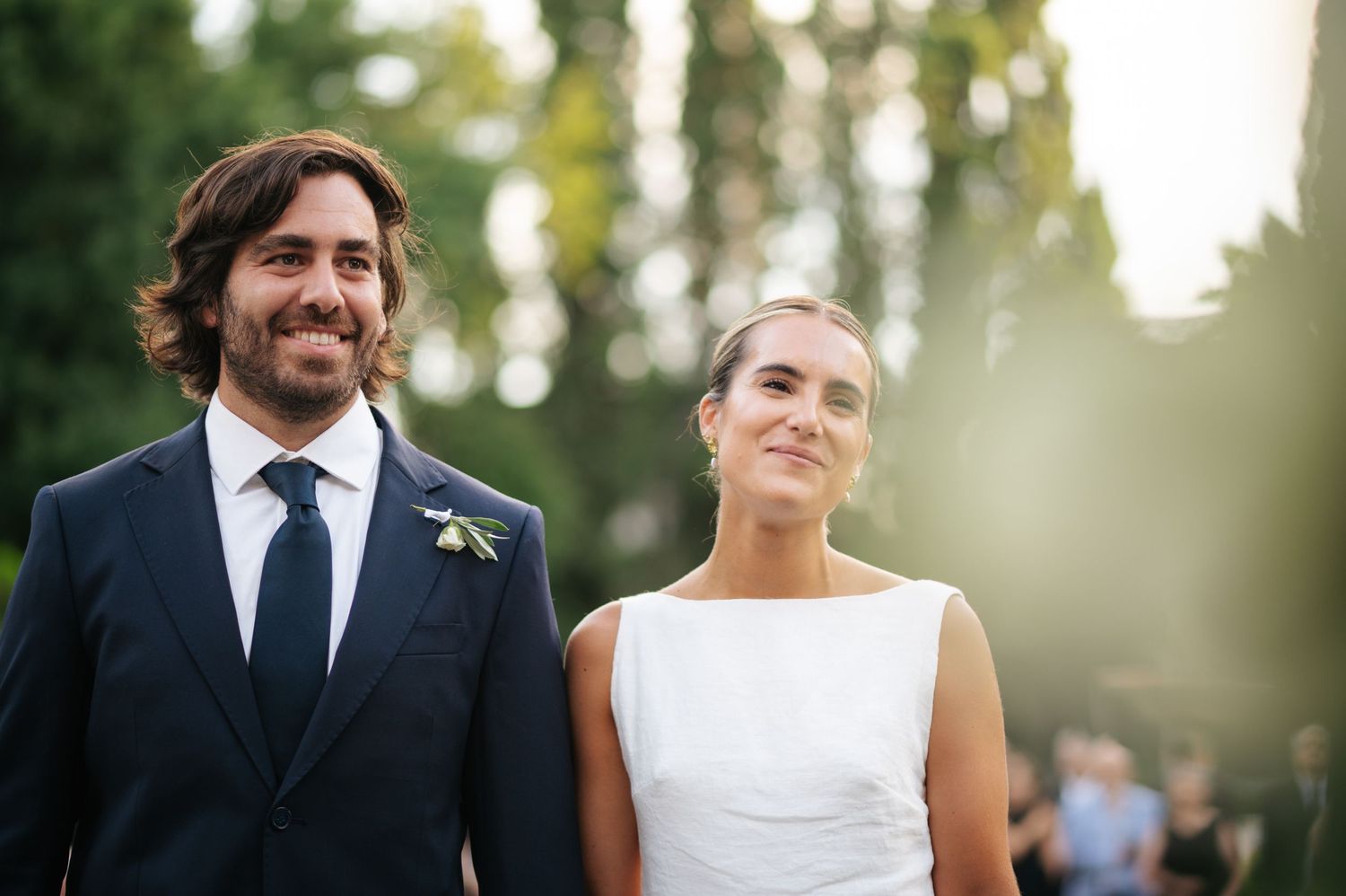 Smiling bride and groom in navy suit with boutonniere walking outdoors at golden hour wedding ceremony.