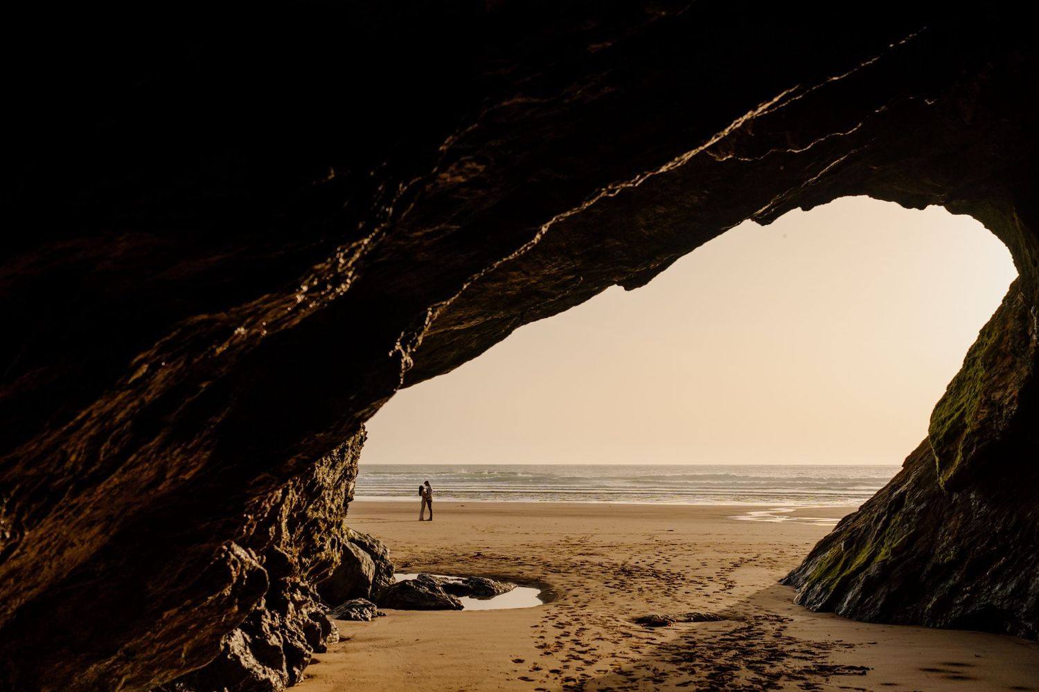 Couple standing on a sandy beach framed by a dramatic sea cave arch with ocean waves in the background.