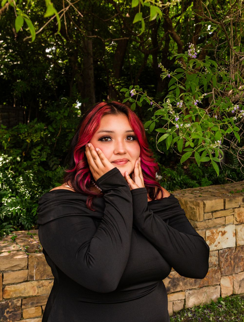 Young woman with red hair in black off-shoulder top poses against stone wall with lush green trees behind her.