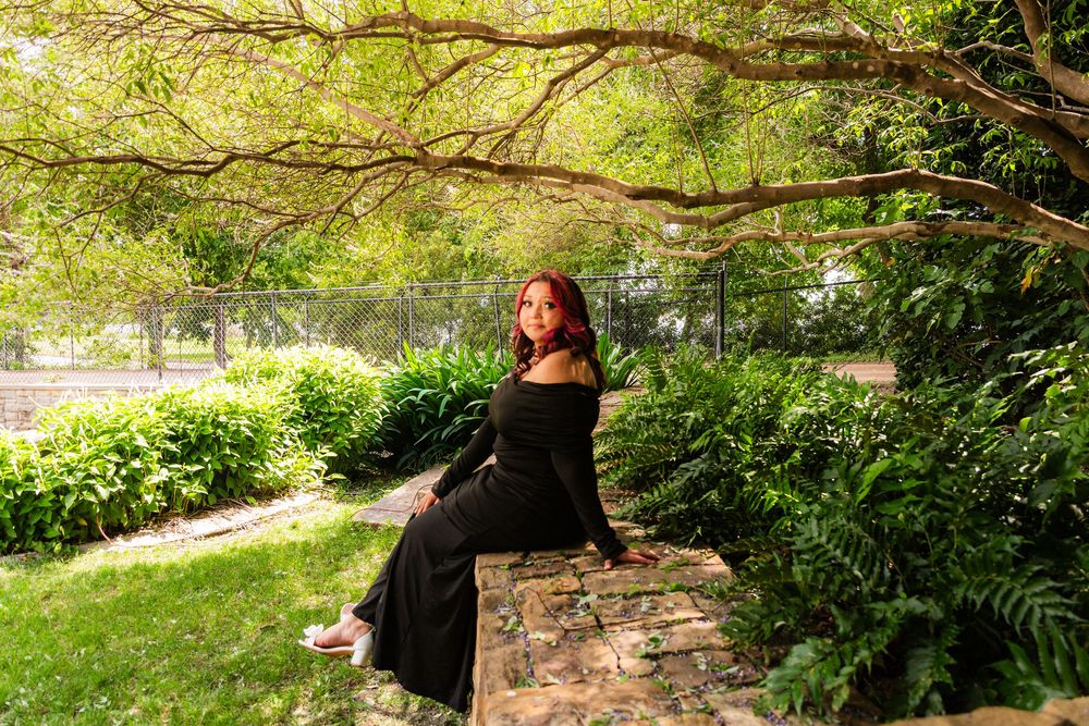 Woman in black dress sitting on stone wall surrounded by lush green garden trees and ferns.