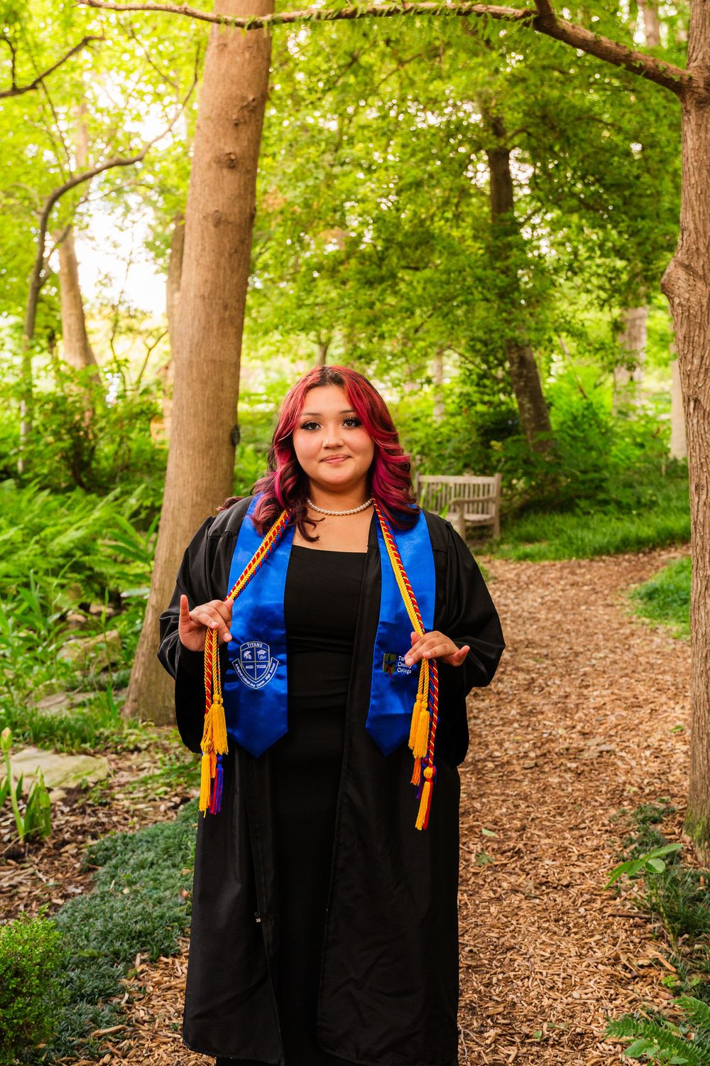 Young woman in black graduation gown with blue stole and gold cords posing in a lush green garden path.