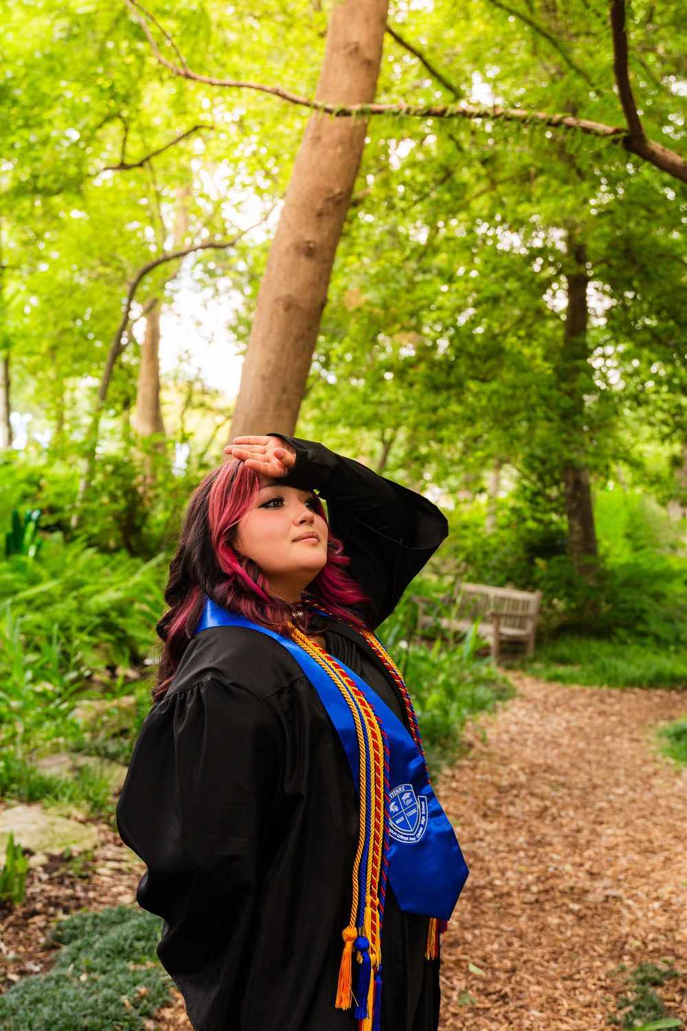 A graduate in black gown with blue and gold stoles poses on a garden path surrounded by lush green trees.