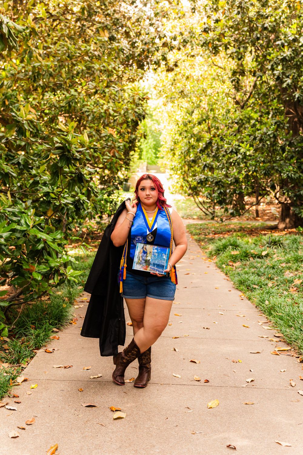 Woman with pink hair in blue top, denim shorts, and boots walks tree-lined path holding black jacket and book.