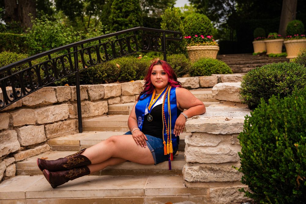 Young woman with red hair in blue graduation dress and cowboy boots poses on stone steps in a garden.