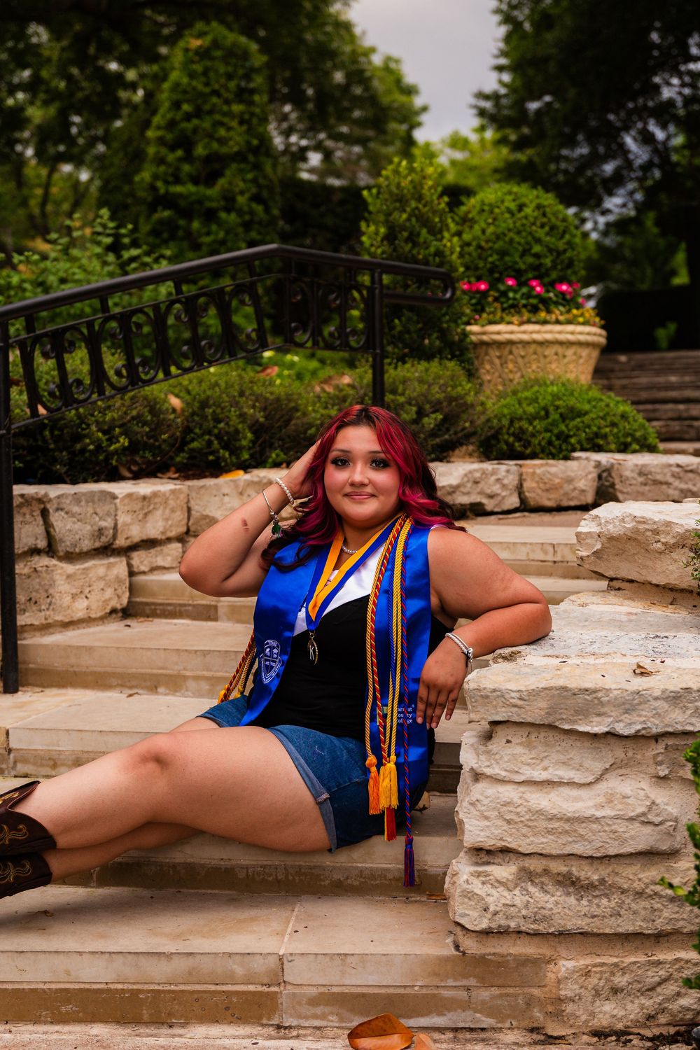 Young woman in blue graduation stole and gold tassels sits on stone steps surrounded by lush greenery.
