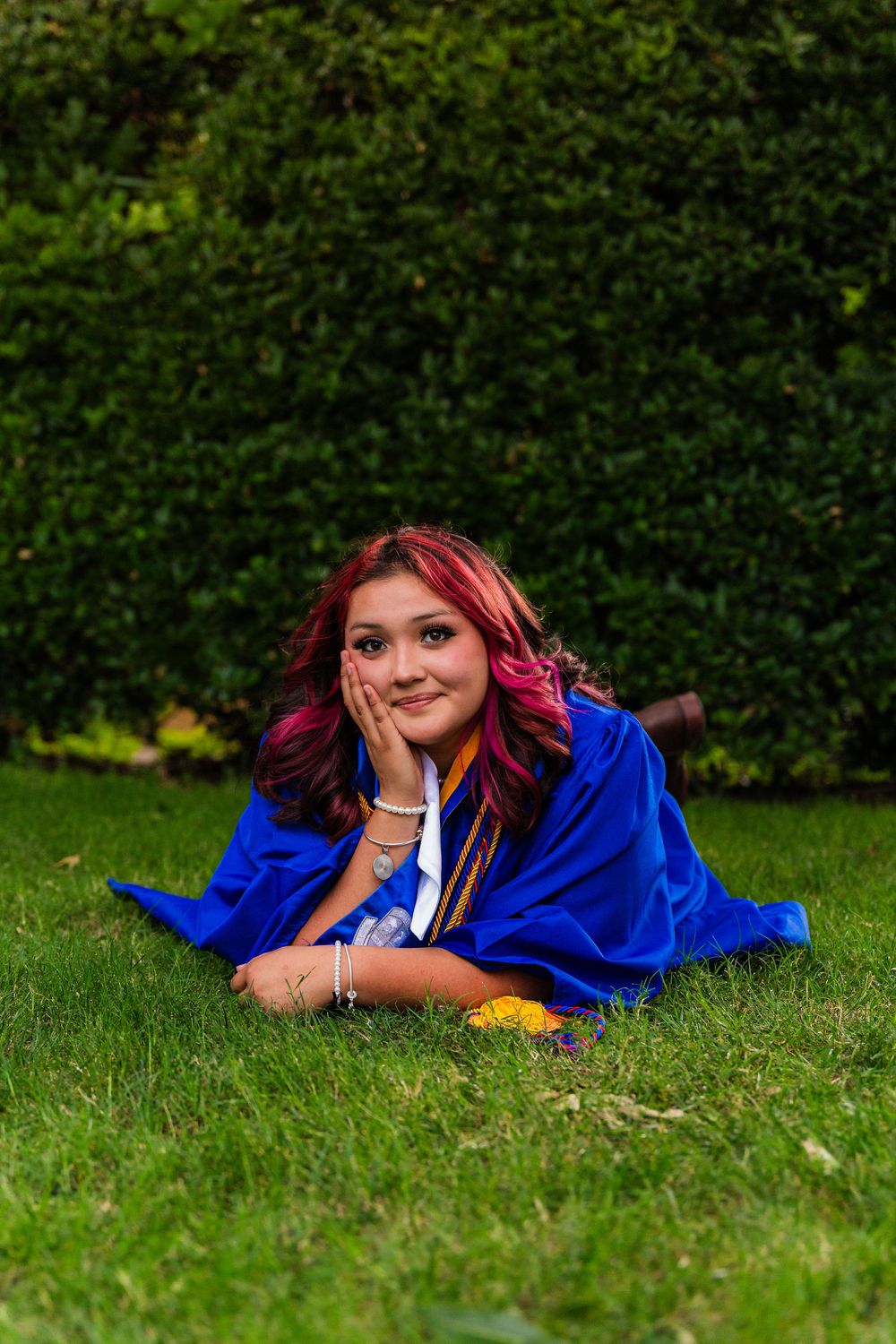 Young woman in blue graduation gown lying on green grass, smiling dreamily with hand on cheek, red highlights in hair.