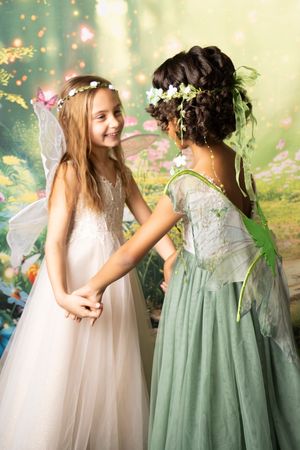 Two girls in fairy costumes with flower crowns and wings hold hands and smile in front of a whimsical, flower-filled woodland backdrop.