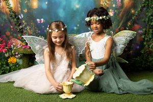 Two young girls in fairy wings and flower crowns play tea party with pastel teapot and teacups on grass in an enchanted garden backdrop.