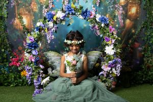 Young girl in a flowing green tulle dress with fairy wings and a flower crown sits inside a floral arch of blue, purple and pink blooms.