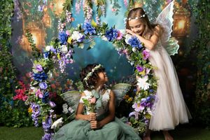 Two girls in fairy costumes with wings pose by a floral swing arch of blue, purple and pink flowers in an enchanted garden photoshoot.