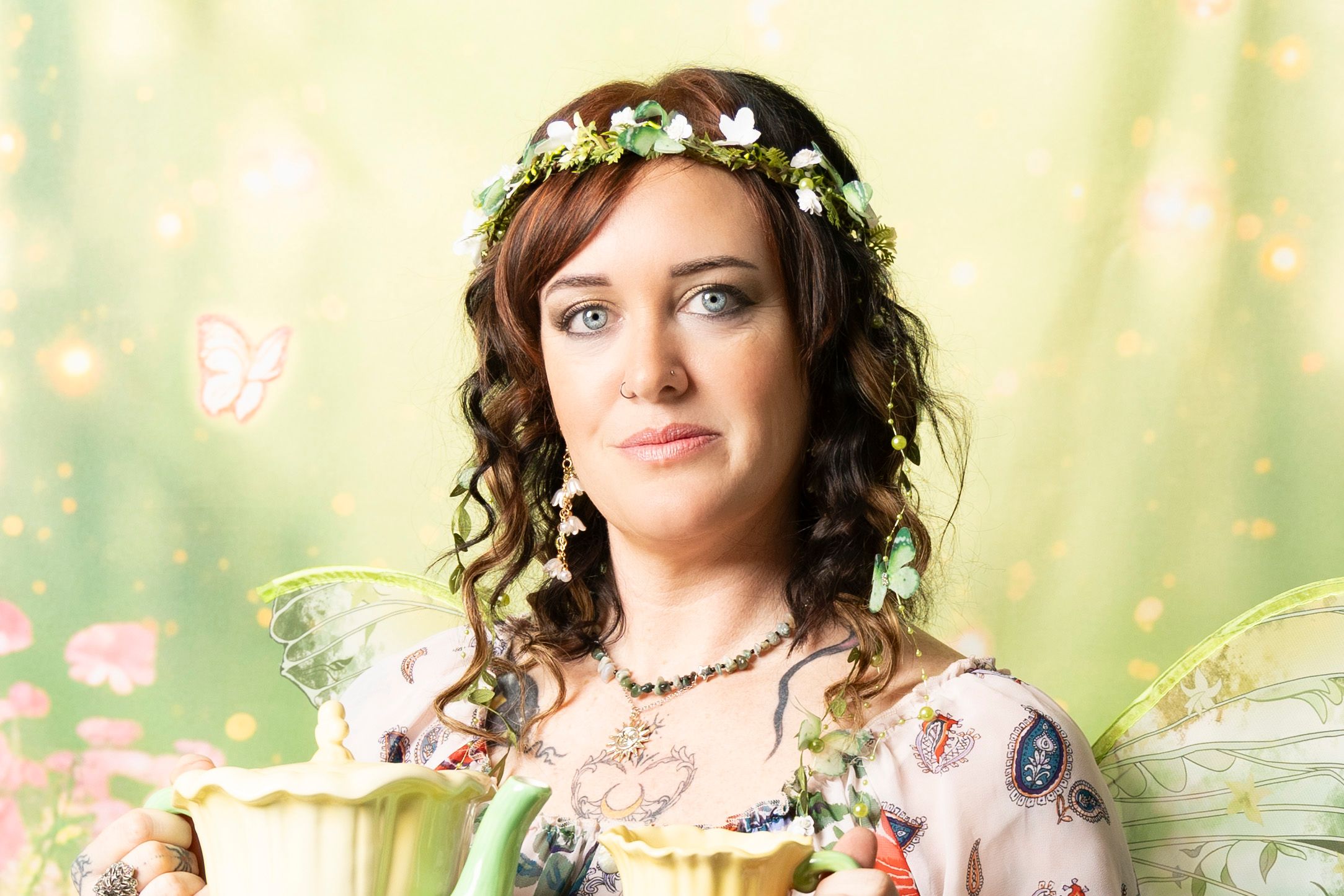 Woman with curly brown hair wearing a floral crown and sheer fairy wings, holding teacup flowers against a soft green garden backdrop.