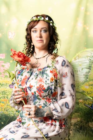 Woman in a floral peasant dress and flower crown holding a red rose, with sheer fairy wings against a whimsical butterfly garden backdrop.