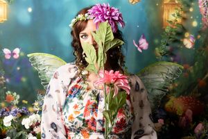 Woman in a floral dress and fairy wings hides half her face behind a green leaf and pink flower in an enchanted garden with butterflies.