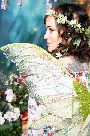 Woman in profile wearing a floral crown and large translucent fairy wings, posed in a whimsical enchanted garden with white flowers.