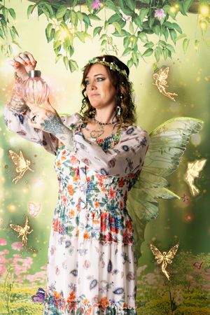 Woman dressed as a fairy with green wings and a flower crown holds a glowing perfume bottle in an enchanted forest with golden fairies.