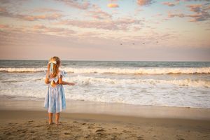A young girl in a blue dress stands on a sandy beach at sunset, gazing at ocean waves under a colorful sky.