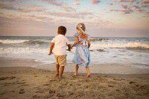 Two young children holding hands on a sandy beach at sunset, waves crashing behind them.
