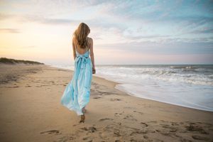 Woman in light blue maxi dress with bow walks barefoot on sandy beach at sunset.