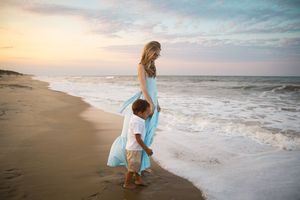 A mother in a blue dress walks with her toddler son along a sandy beach at sunset.