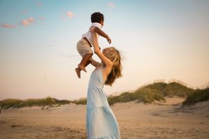 A mother in a white dress lifts her baby joyfully on a sandy beach at sunset.