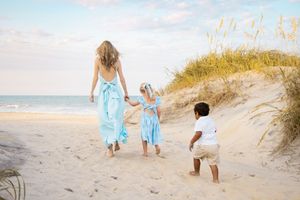 Woman in blue dress walks with two young children on a sandy beach toward the ocean on a sunny day.